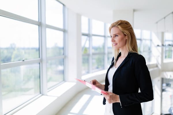 Businesswoman standing by a large window holding a folder, looking thoughtfully ahead.